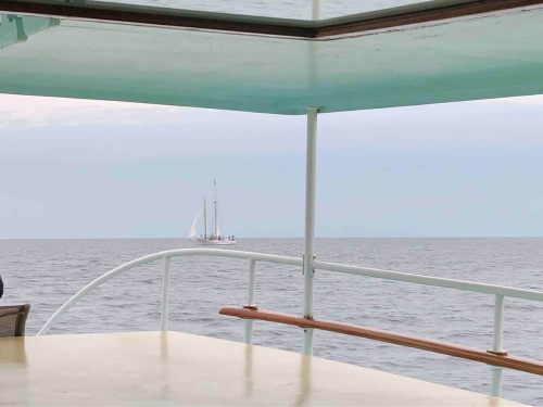 A view from a boat's deck overlooking the ocean, with a sailboat in the distance against a cloudy sky.