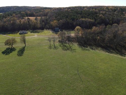Aerial view of a green field with scattered trees and two houses in a rural landscape surrounded by wooded hills.