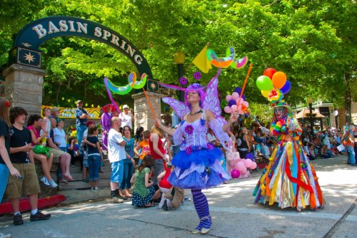 A vibrant parade scene with colorful costumes, balloons, and a crowd enjoying the festivities under green trees.