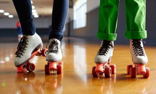 Two pairs of roller skates on a wooden floor, one in black pants and the other in bright green pants.