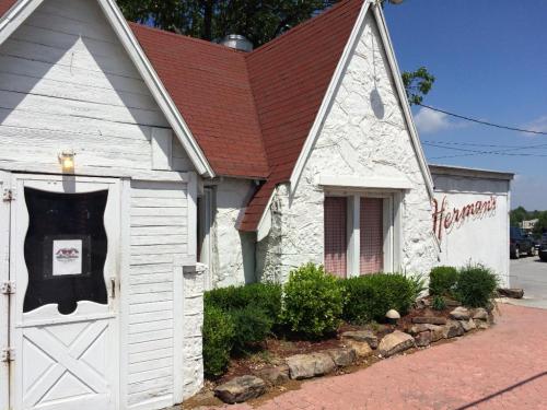 A quaint white building with a red roof and "Herman's" sign, surrounded by greenery and a stone border.