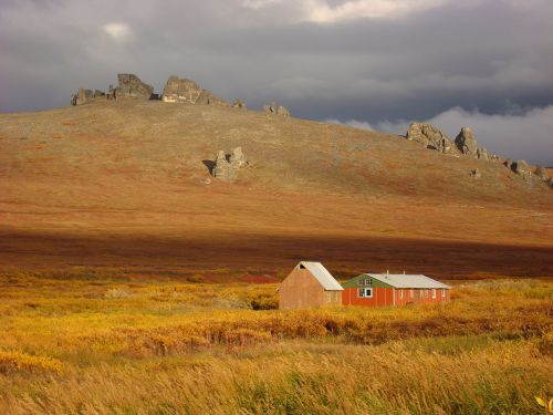 A red and green house sits in a colorful field, with rocky hills and dark clouds in the background.