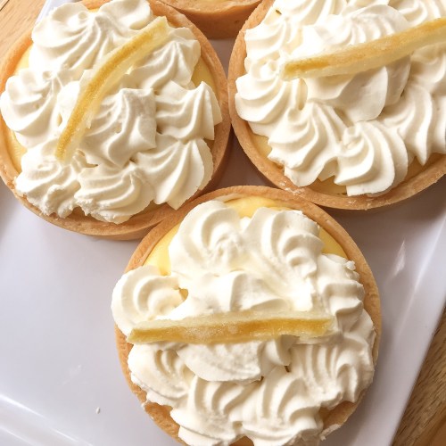 Four mini lemon tarts topped with whipped cream and lemon slices, arranged on a white plate.