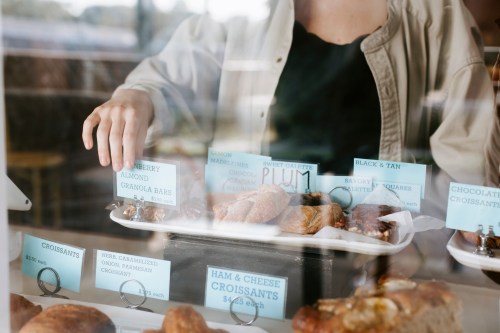 A hand reaches for pastries behind a glass display, featuring various labeled baked goods like croissants and granola bars.
