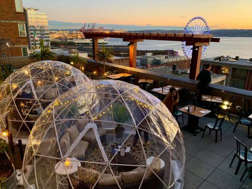 Rooftop dining with transparent igloos, city skyline, and a Ferris wheel at sunset over the water.