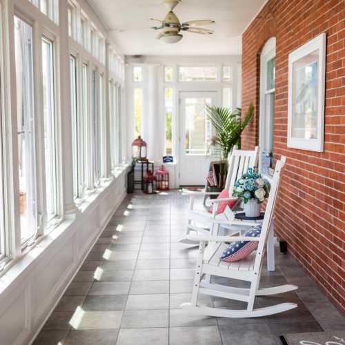 A bright sunroom with rocking chairs, potted plants, and patriotic decor, featuring large windows and tiled flooring.