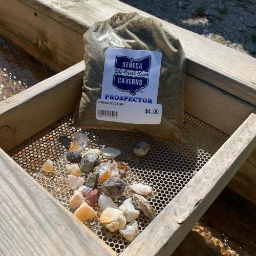 A bag of "Prospector" sand sits on a wooden surface next to various colorful gemstones and minerals.