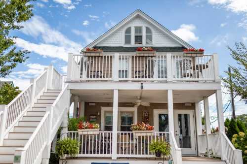 A charming two-story house with a large front porch, white railings, and colorful flower pots under a blue sky.