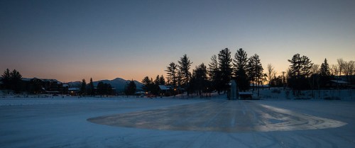 A serene winter landscape at dusk, featuring a frozen lake surrounded by trees and distant mountains.