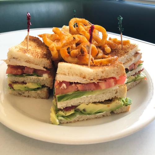 A stacked sandwich with layers of lettuce, tomato, and avocado, topped with curly fries, served on a white plate.