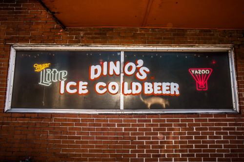 Sign for Dino's Ice Cold Beer featuring Miller Lite and Yazoo logos, set against a brick wall.