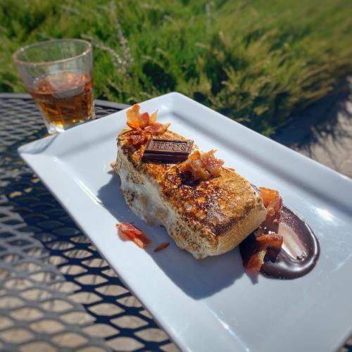 A gourmet s'more with toasted marshmallow, chocolate, and bacon bits, served on a white plate with a drink in the background.