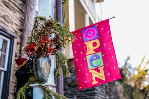 A colorful "OPEN" banner hangs beside a vase of greenery and berries, set against a rustic building backdrop.