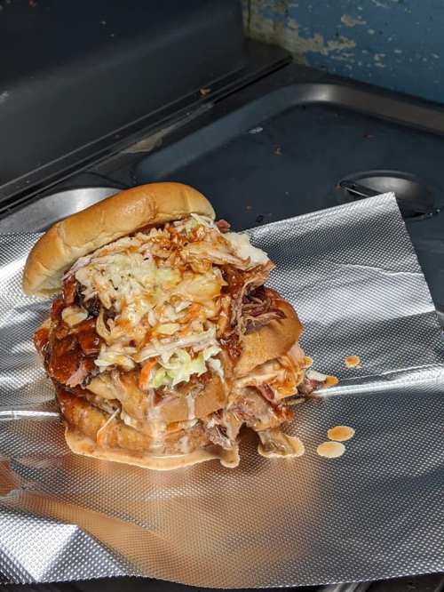 A messy burger stacked with pulled pork, cheese, and coleslaw, resting on foil in a kitchen setting.