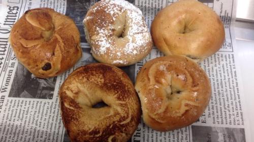 A variety of five bagels on a newspaper, including plain, cinnamon, and powdered sugar varieties.