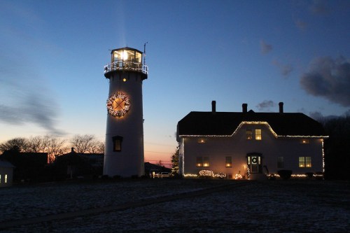 A lighthouse and a house decorated with lights at dusk, with a colorful sky in the background.