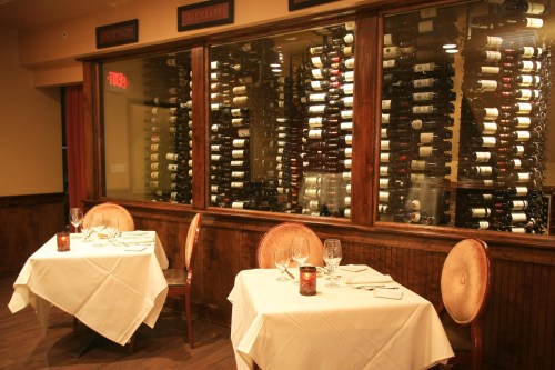 Two elegantly set tables with white tablecloths in a restaurant, featuring a large wine display in the background.