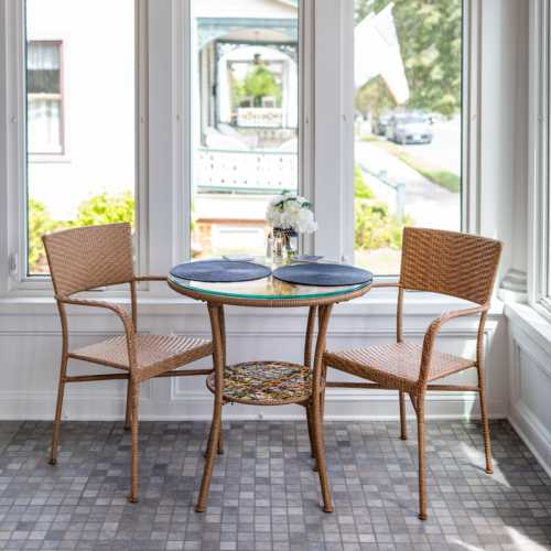 A cozy dining area with a round table and two wicker chairs, surrounded by large windows and a view of greenery outside.