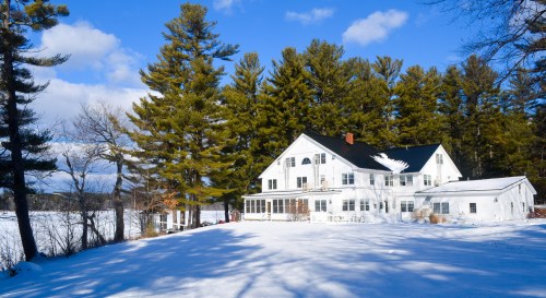 A white house surrounded by snow and tall pine trees under a blue sky with fluffy clouds.