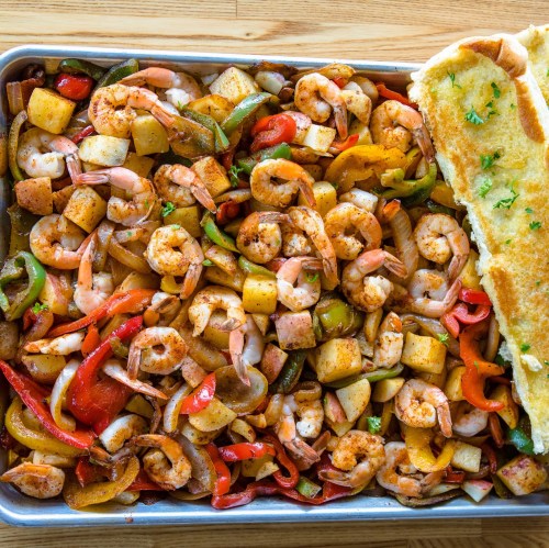 A tray of seasoned shrimp, colorful bell peppers, onions, and potatoes, served with a piece of toasted bread.