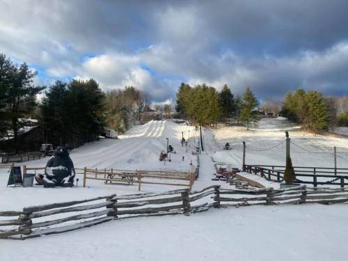 Snow-covered landscape with a sledding hill, trees, and a wooden fence under a cloudy sky.