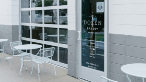 Exterior of Dozen Bakery with a glass door, small tables outside, and a sign displaying hours and location.