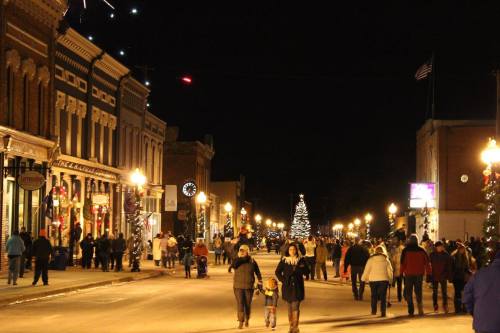 A festive street scene at night, with people walking past decorated shops and a Christmas tree in the distance.
