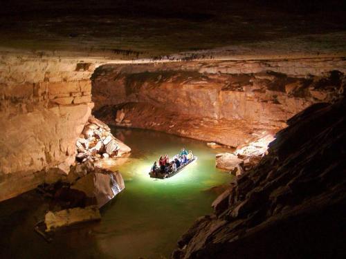 A small boat with people navigates through a dimly lit cave with rocky walls and green water.