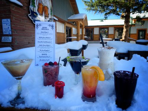 A variety of colorful cocktails and drinks displayed on snow, with a menu in the background and a rustic building.