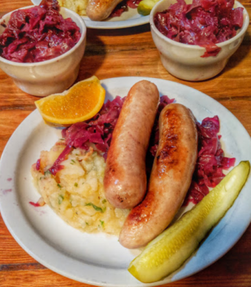 A plate with two sausages, red cabbage, mashed potatoes, a pickle, and an orange slice, served with two small bowls of cabbage.
