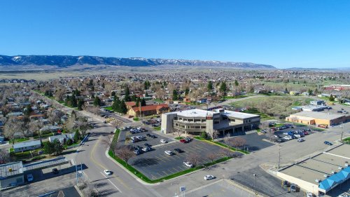 Aerial view of a small town with buildings, parking lots, and mountains in the background under a clear blue sky.
