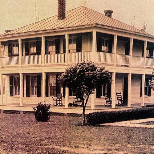 A historic two-story house with a wraparound porch, surrounded by greenery and a well-maintained lawn.