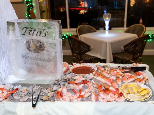 A seafood display featuring shrimp, oysters, and lemon, with an ice sculpture labeled "Tito's Handmade" in the background.