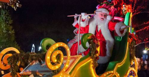 Santa Claus in a festive sleigh, waving joyfully during a nighttime holiday parade, surrounded by colorful lights.