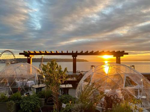 Sunset over a waterfront restaurant with clear dining domes and a view of a Ferris wheel in the distance.