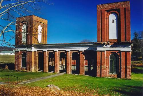 Ruins of a brick building with two towers, surrounded by grass and trees under a clear blue sky.