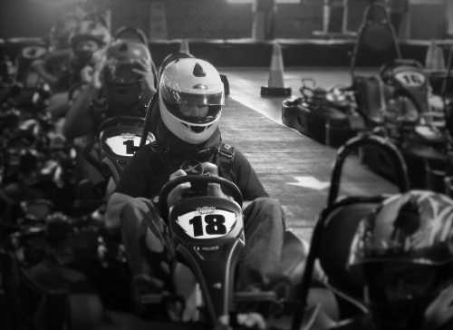 A go-kart racer in a helmet sits in a line of karts, ready to race on an indoor track.