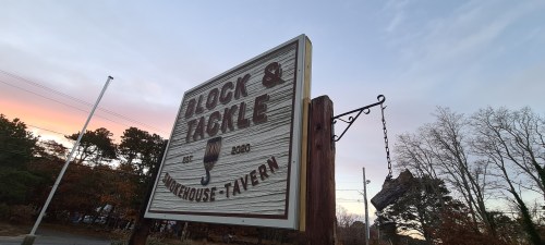 Sign for "Block & Tackle" restaurant and tavern, featuring a fishing motif, against a colorful sunset sky.