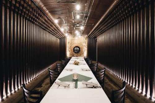 A long, narrow dining room with wooden walls, a central table set for a meal, and soft lighting.