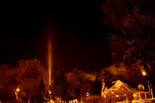 A night scene featuring a bright vertical light beam above trees and a house, with a dark sky and streetlights.