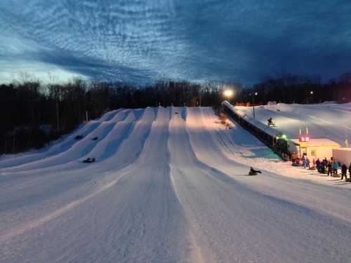 Snow-covered tubing hill with multiple lanes, illuminated by lights, under a twilight sky. People are enjoying the slopes.