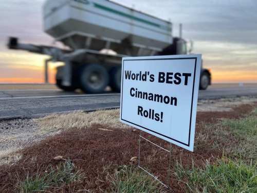 A sign reading "World's BEST Cinnamon Rolls!" stands by the roadside with a blurred truck in the background at sunset.