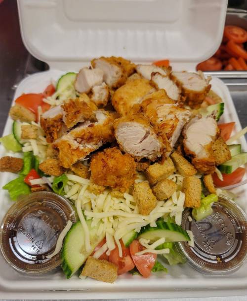 A salad topped with sliced fried chicken, croutons, cheese, and served with two dressing containers.
