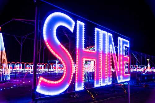 Brightly lit sign reading "SHINE" in colorful lights against a dark background, surrounded by festive decorations.