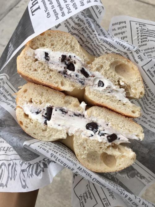 A bagel cut in half, filled with cream cheese and crushed Oreo cookies, served on newspaper print.