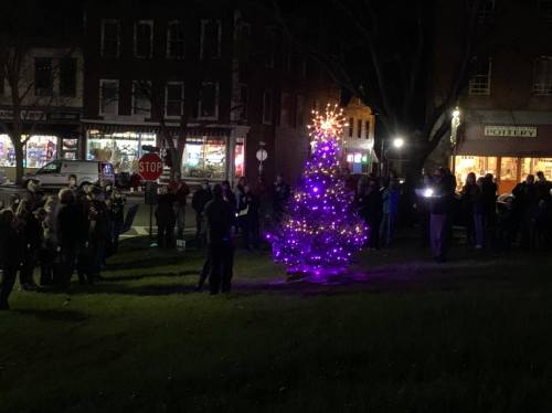 A crowd gathers around a brightly lit Christmas tree in a festive nighttime setting.