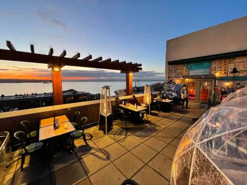 A rooftop dining area with tables, heaters, and a sunset view over the water, featuring a mural on the wall.