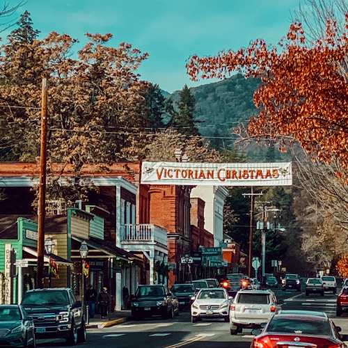 A charming street scene featuring Victorian-style buildings and festive decorations, surrounded by trees and mountains.
