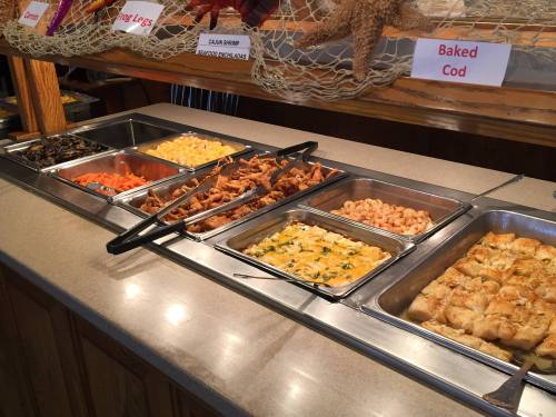 A buffet display with various seafood dishes, including shrimp, baked cod, and sides like carrots and bread rolls.