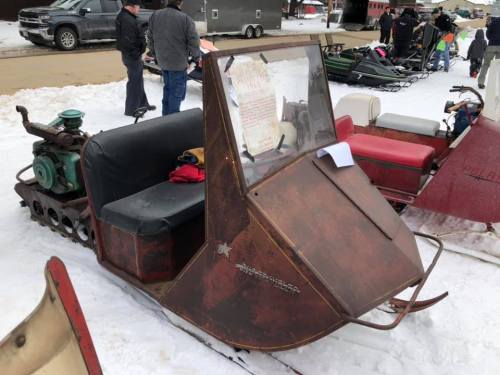 A vintage snowmobile with a metal body and a green engine, parked on snow with people in the background.
